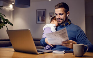 Man looking at computer with a document in one hand and a baby sleeping on his shoulder. Presumably shopping benefits coverage.